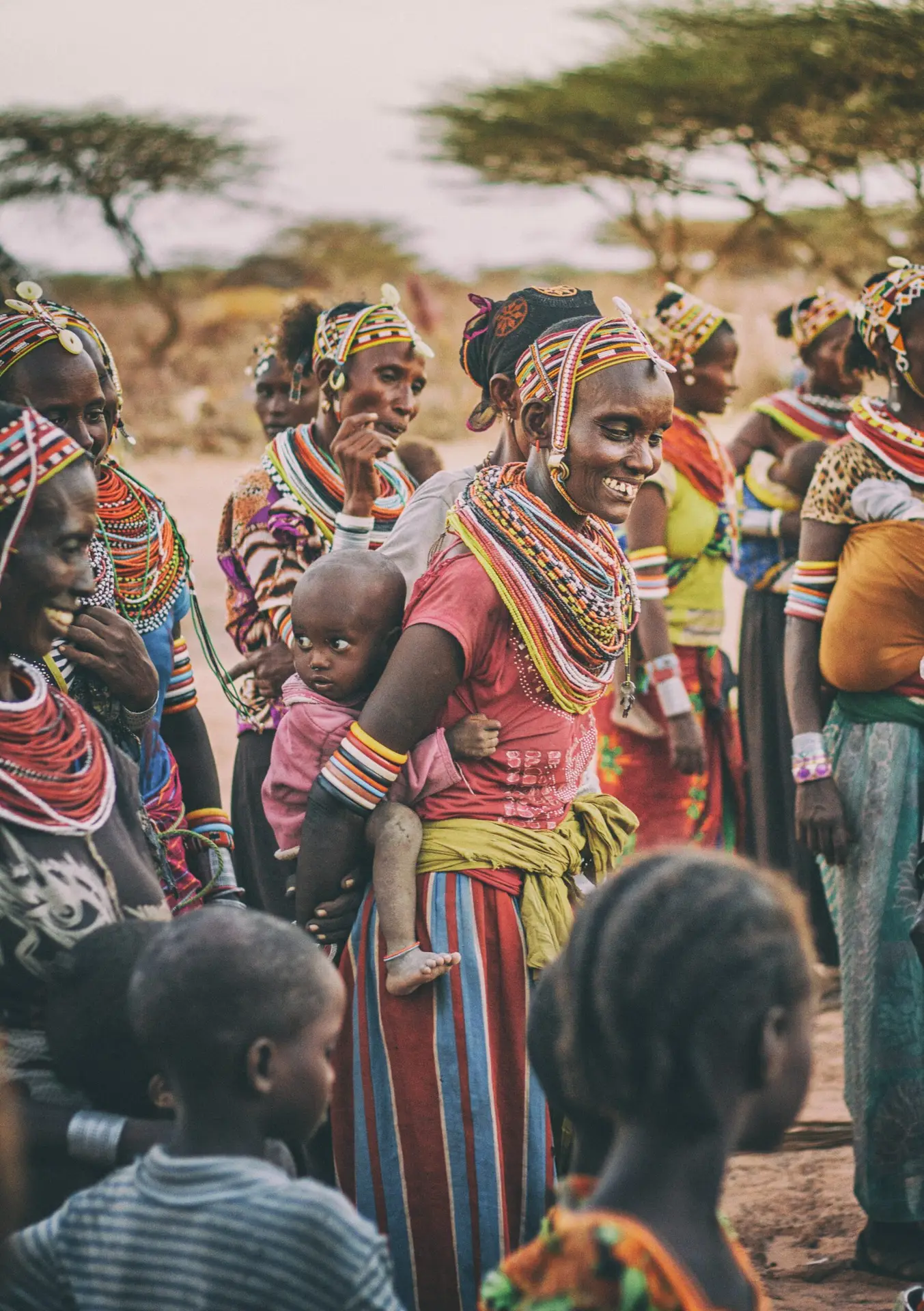Femme kenyane souriante portant son enfant, entourée de femmes en tenues traditionnelles. Voyage d’aventure sur mesure conçu par Trace ta Roots